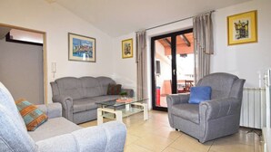 A living room with a grey sofa, a grey armchair, and a glass coffee table. There are two framed pictures on the wall, a sliding glass door leading to an outdoor area, and a radiator on the right. - Apartments Edo (Stinjan)