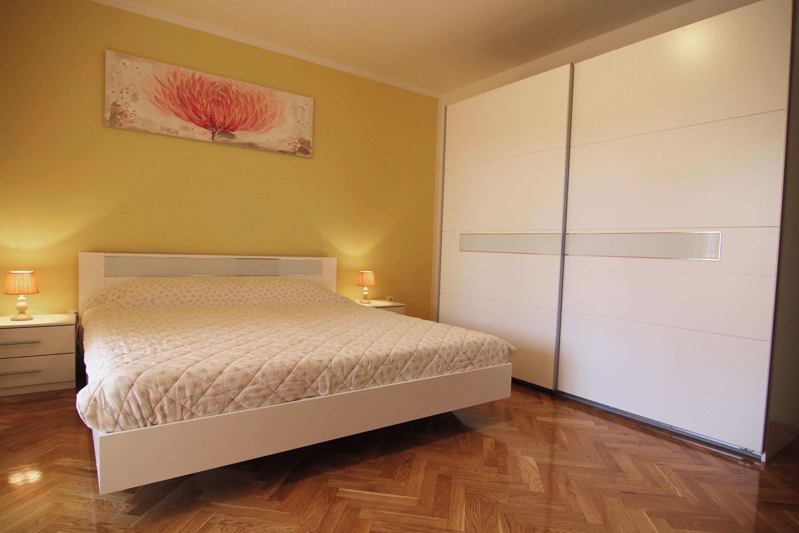 A bedroom with a bed, bedside lamps, a nightstand, a wardrobe, and a floral painting on the wall.