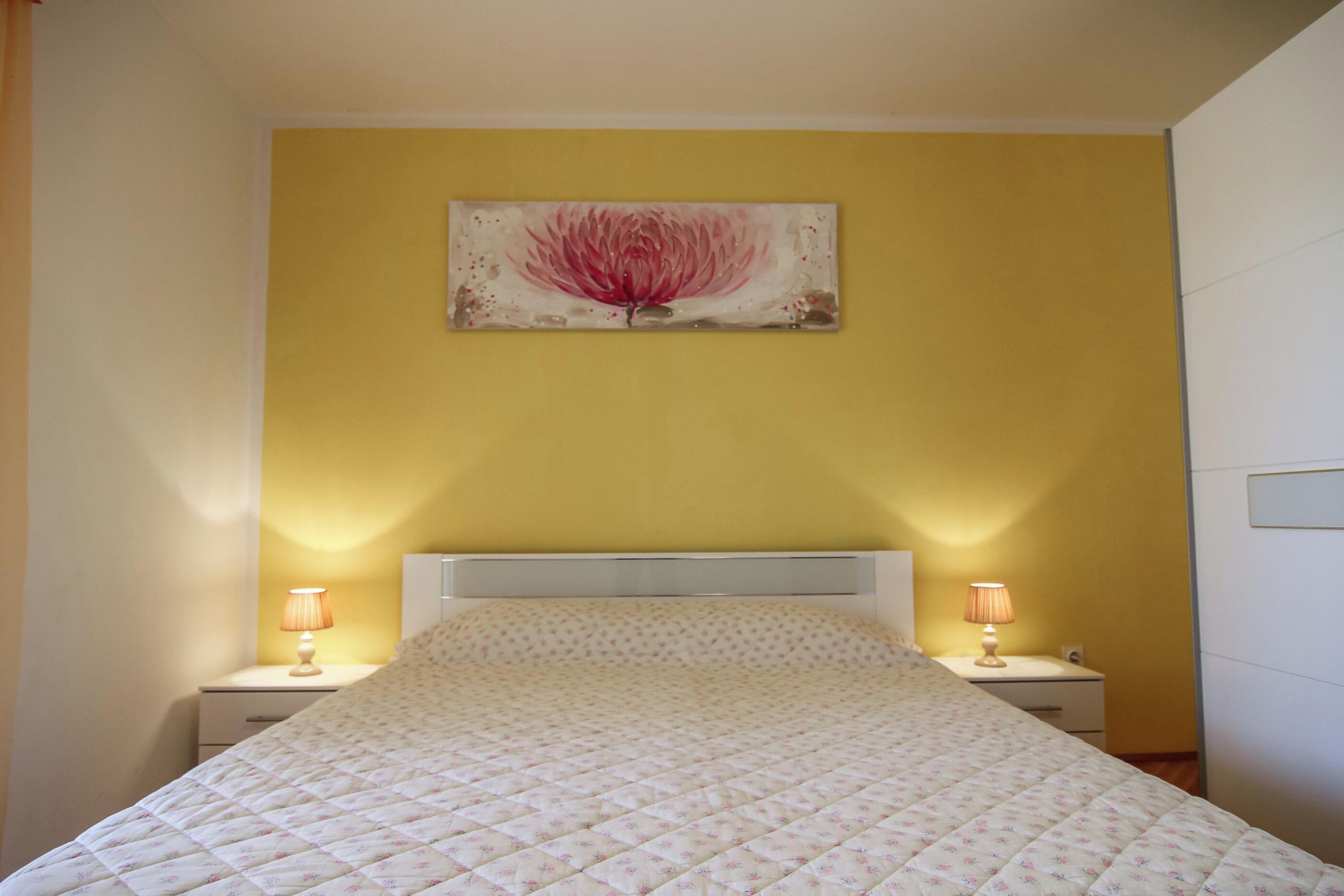 A bedroom with a bed, two bedside tables with lamps, a wall-mounted artwork, and a white headboard.