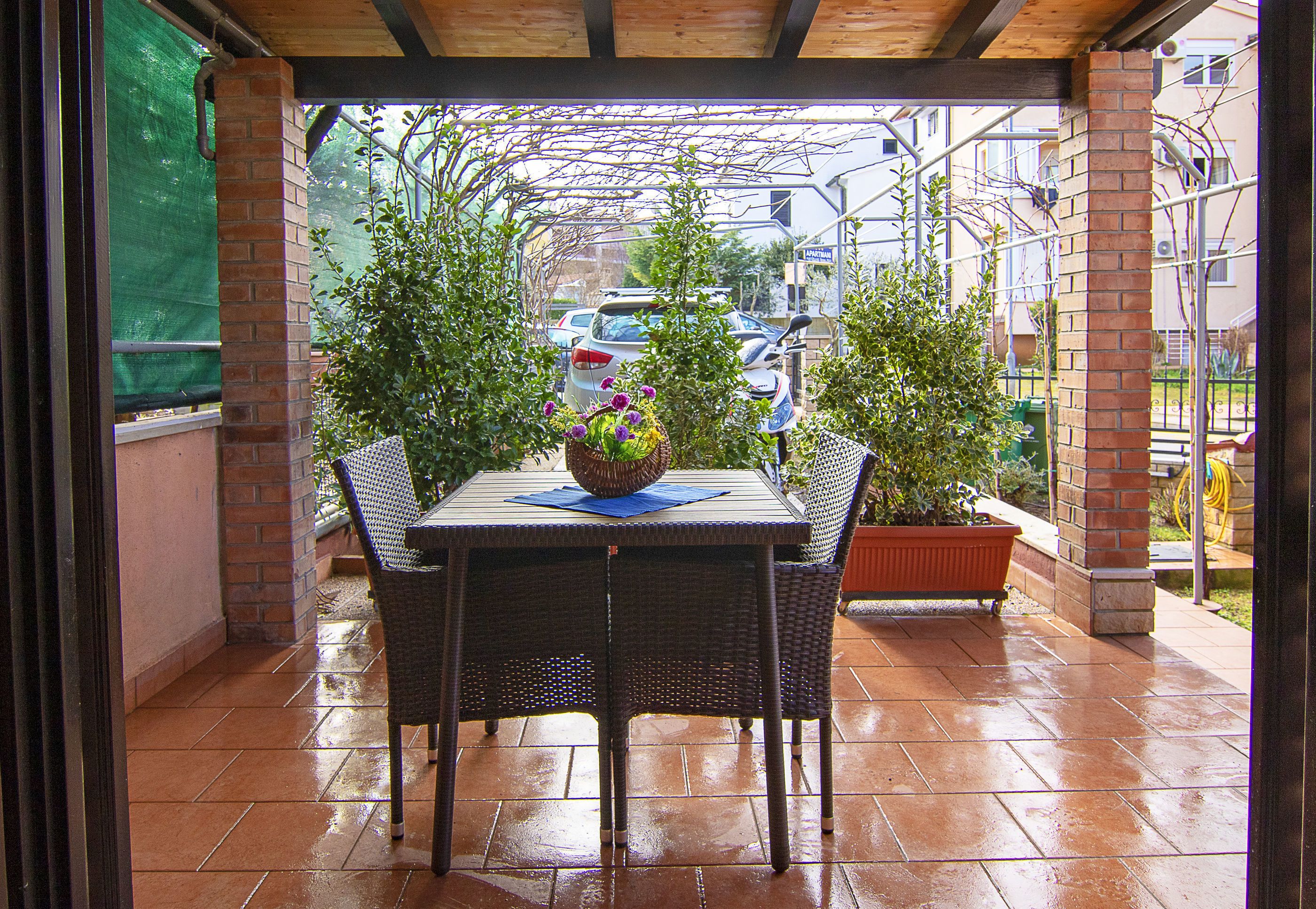 A covered patio with a table and chairs, surrounded by potted plants and a view of parked cars and buildings in the background.