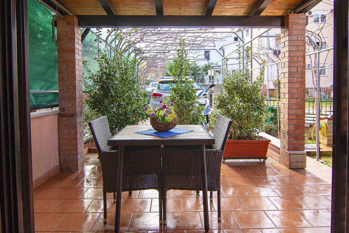 A covered patio with a table and chairs, surrounded by potted plants and a view of parked cars and buildings in the background.