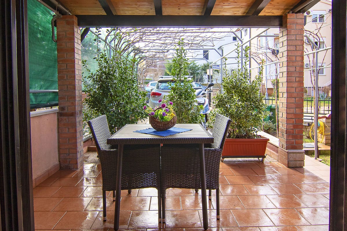 A covered patio with a table and chairs, surrounded by potted plants and a view of parked cars and buildings in the background.
