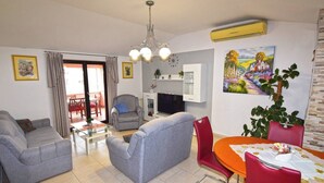 A living room with grey sofas, a dining table with red chairs, a TV, and a painting on the wall. - Apartments Edo (Stinjan)