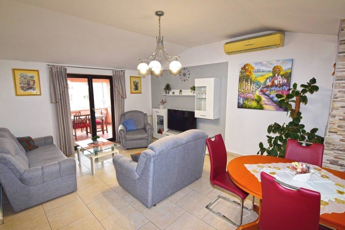 A living room with grey sofas, a dining table with red chairs, a TV, and a fireplace.