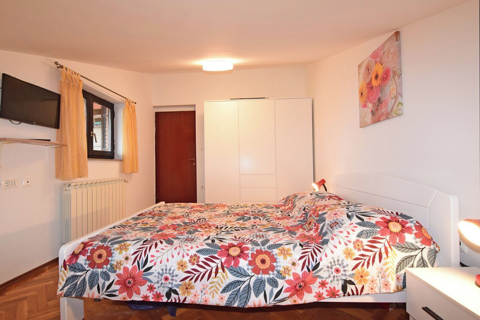 A bedroom with a bed, a bedside lamp, a radiator, a TV mounted on the wall, and a door.