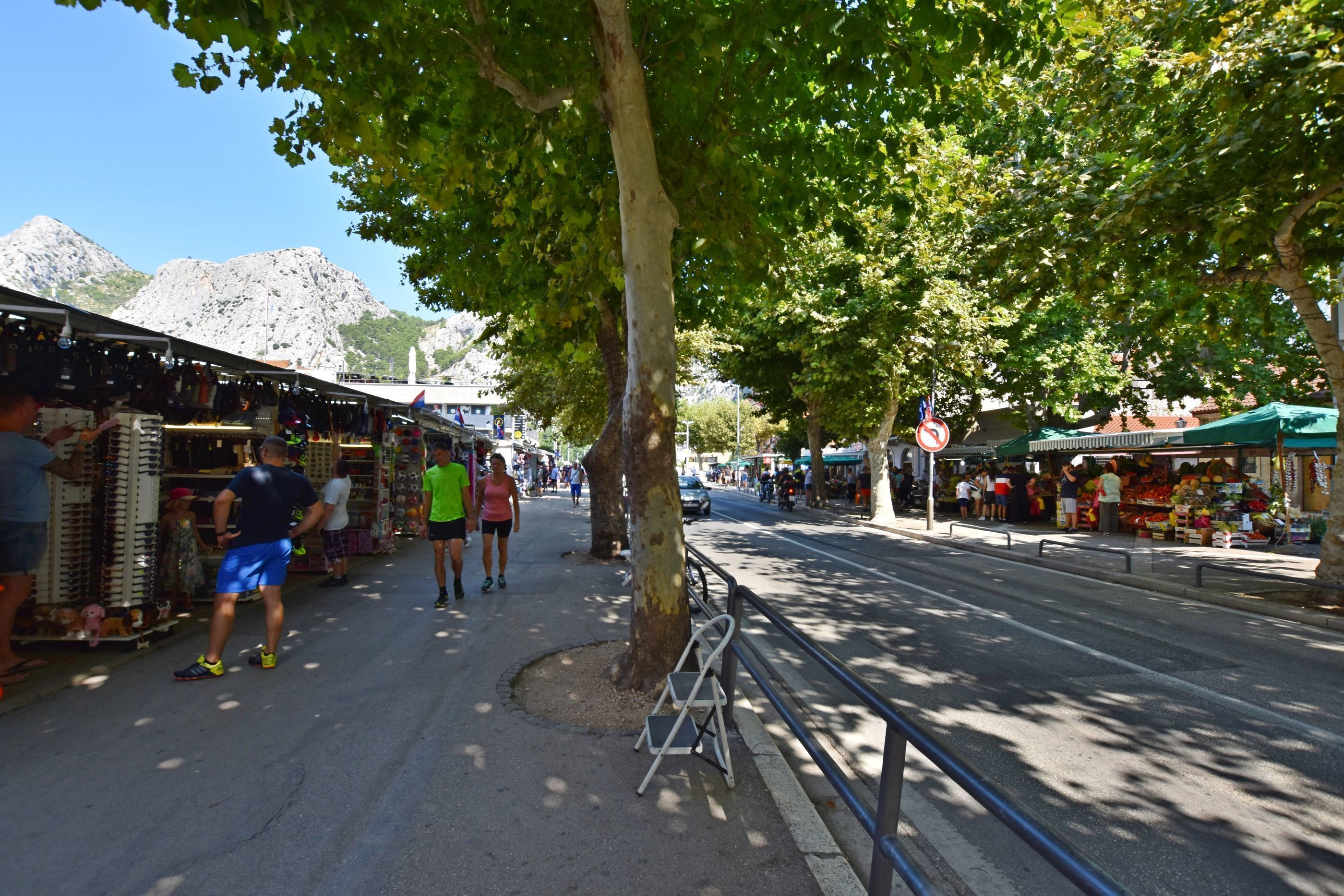 Een straatmarkt met verkopers en bezoekers, bomen langs de stoep en bergen op de achtergrond.