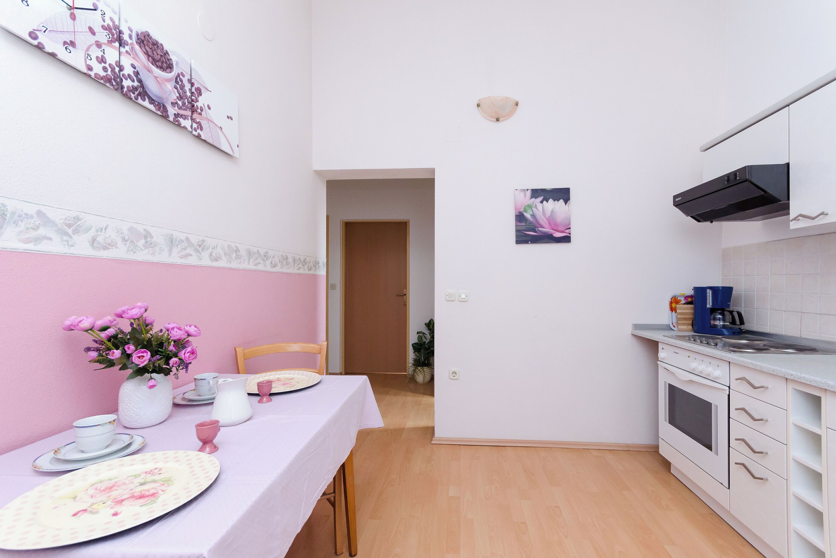 A kitchen with white cabinets, a sink, and a stove. There is a dining table with pink flowers and plates.