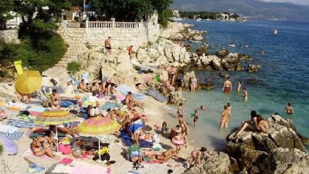 Une plage bondée où les gens se prélassent au soleil et se baignent, un mur de pierre et une mer d’un bleu limpide.