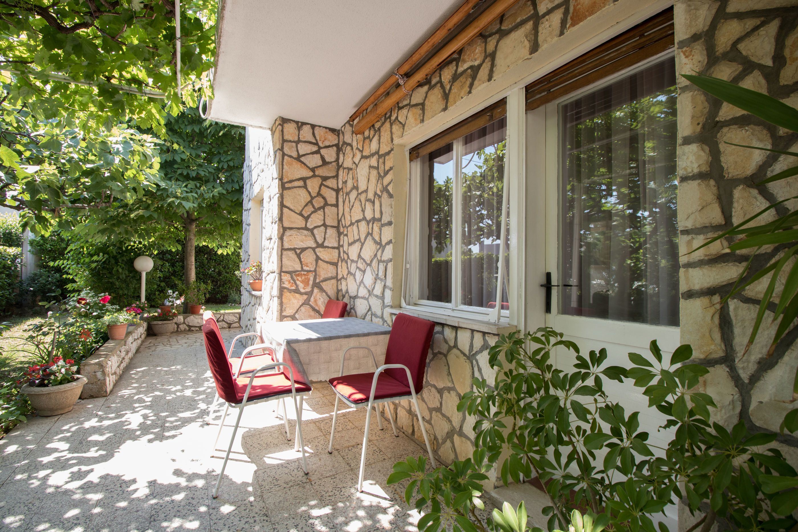 Une façade en pierre avec une fenêtre, une terrasse avec une table et des chaises, et des plantes en pot.