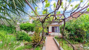 A garden pathway leading to a house with a red brick entrance, surrounded by various plants and trees. - Apartments Tugomila (Barbat)