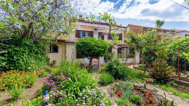 A residential garden with a variety of plants and flowers, a small gnome statue, and a two-story house with a balcony. - Apartments Tugomila (Barbat)