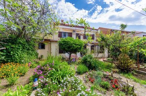 A residential garden with a variety of plants and flowers, a small gnome statue, and a two-story house with a balcony.
