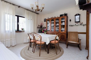 Une salle à manger avec une table ronde, six chaises en bois, un lustre, un buffet aux portes vitrées et une fenêtre avec des rideaux.