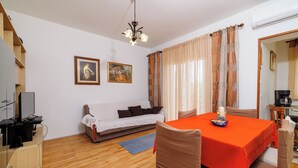 A room with a wooden floor, a white sofa with a blanket, a dining table with a red tablecloth, and two framed pictures on the wall.