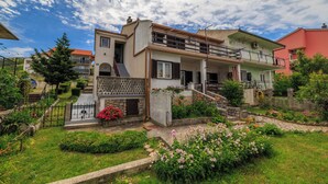 A two-story house with a balcony, a garden with flowers, and a clear sky.