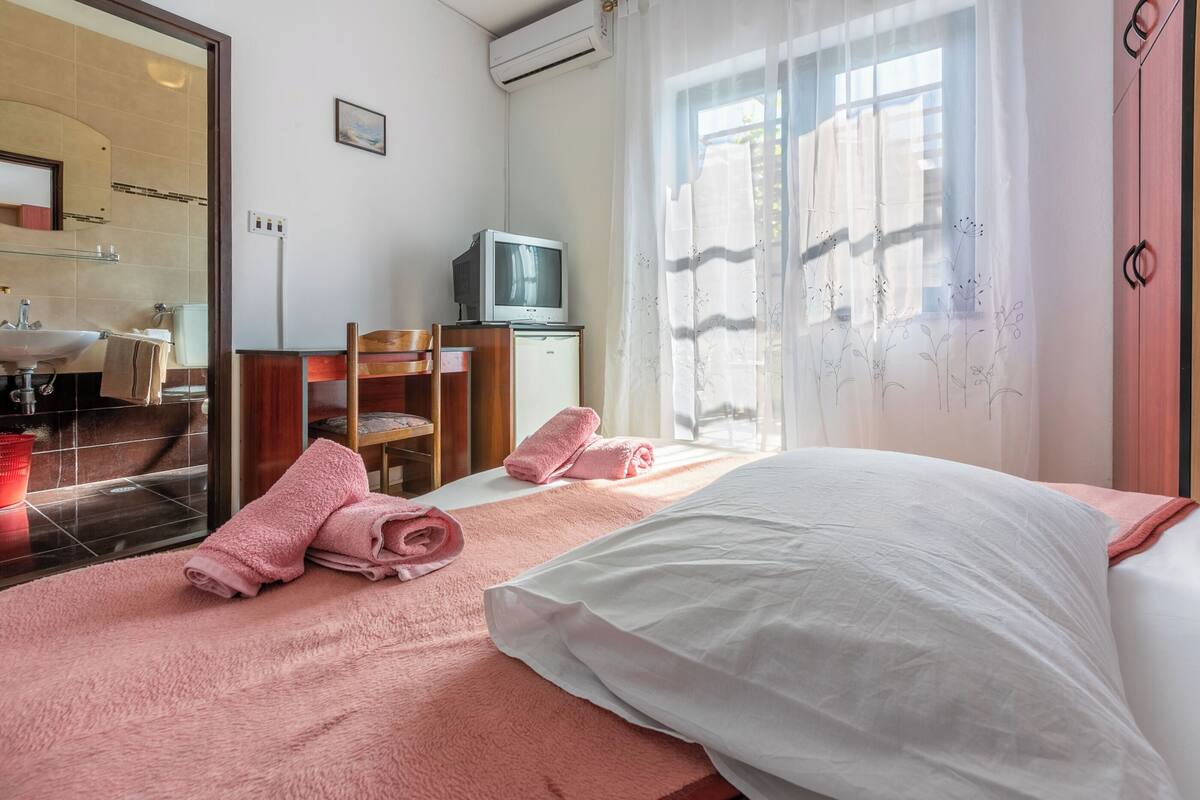 Ein Hotelzimmer mit Bett, Fernseher, Schreibtisch und angrenzendem Badezimmer, durch eine Glastür sichtbar.