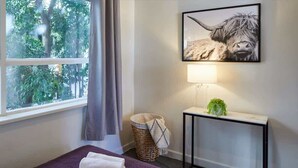 Una habitación de hotel con una cama, una ventana con cortinas, una lámpara de pared, un cuadro enmarcado de un yak y una planta pequeña sobre una mesita consola.