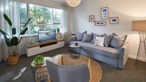 Una sala de estar moderna con un sofá gris, un televisor sobre un mueble bajo, una mesa de centro redonda y un ventanal con cortinas.