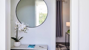 Una habitación con un espejo redondo en la pared, una pequeña consola blanca con un libro y una orquídea en maceta, y una cortina gris al fondo.