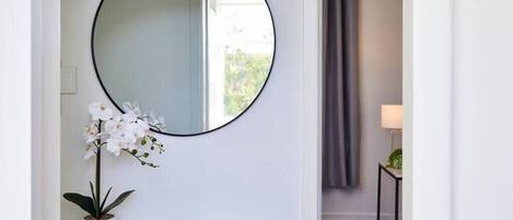 Une pièce avec un grand miroir rond au mur, une petite console blanche sur laquelle sont posés un livre et une orchidée en pot, et un rideau gris en arrière-plan.
