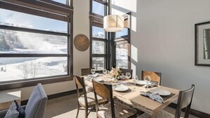 Un comedor con una mesa preparada para cuatro, ventanas amplias con vista a un paisaje nevado, una lámpara fijada a la pared y un cuadro enmarcado.