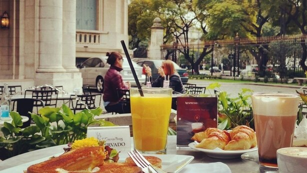 Ein gedeckter Tisch mit Frühstücksangeboten, darunter Croissants, ein Sandwich und ein Getränk, mit Stadtansicht und Bäumen im Hintergrund.