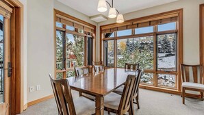 Uma sala de jantar ampla com uma grande mesa de madeira e cadeiras, janelas grandes com vista para uma paisagem nevada e um lustre.