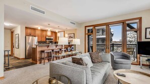 A modern living room with a grey sofa, a coffee table, and a TV. There is a kitchen area with wooden cabinets and a dining table. - Capitol Peak Lodge 3210 (Snowmass Village)