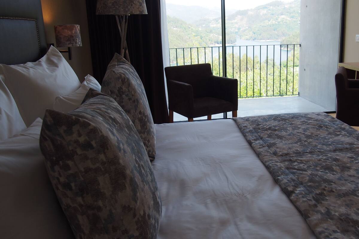 Un dormitorio con una cama grande, una silla y un escritorio. Se ve un balcón con barandilla.