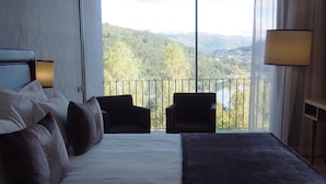 Habitació d'hotel amb un llit gran, dues butaques, vista a una zona boscosa i un balcó amb barana.