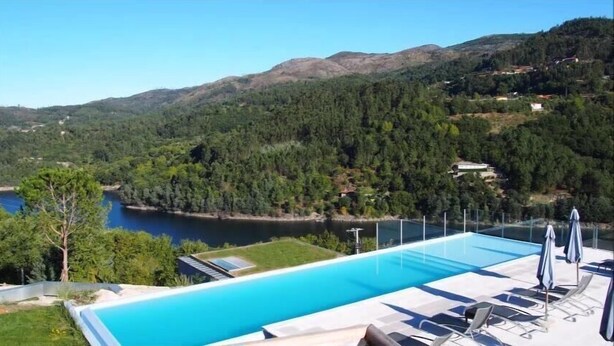 Una piscina amb gandules i ombrel·les, en un entorn de paisatge muntanyós i una massa d'aigua.