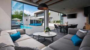 Una sala de estar moderna con vista a una piscina, sofás grises con cojines azules, una mesa de centro redonda y un comedor con televisor.