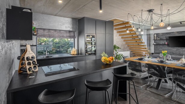 Una cocina moderna con una isla central, taburetes negros, una escalera de madera y un comedor con una mesa dispuesta para dos.