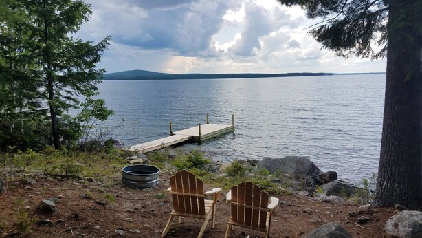 Vista de un lago con un muelle que se adentra en el agua, rodeado de árboles y rocas. Dos sillas Adirondack están ubicadas en la orilla.