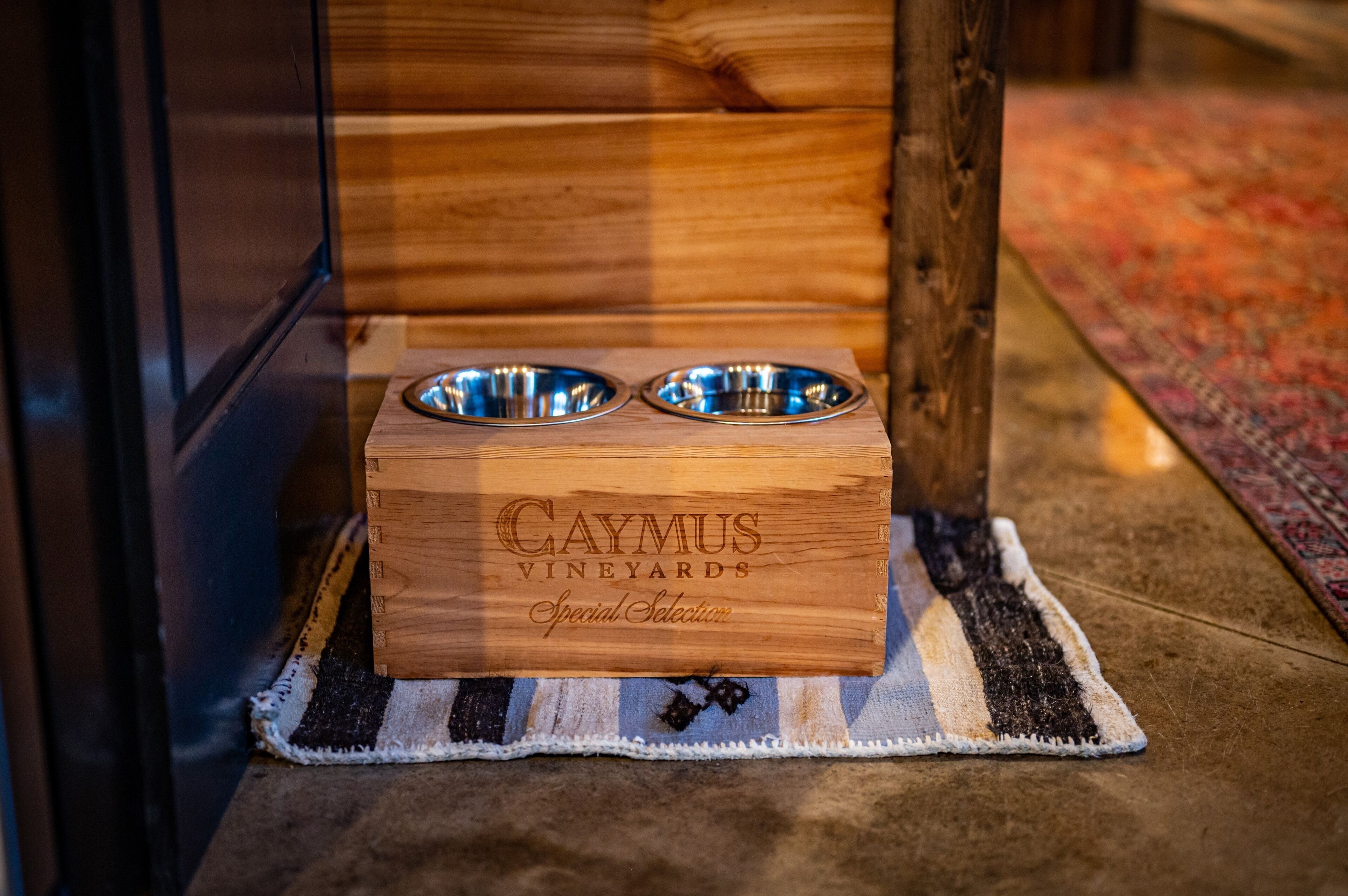 Una caixa de fusta amb la inscripció "CAYMUS VINEYARDS Special Selection" i dos bols metàl·lics a sobre, sobre una catifa amb ratlles.