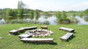 A lakeside setting with a fire pit surrounded by stones and logs, grassy areas, and trees in the background. - Nice apartment in Borcz with kitchen (Borcz)