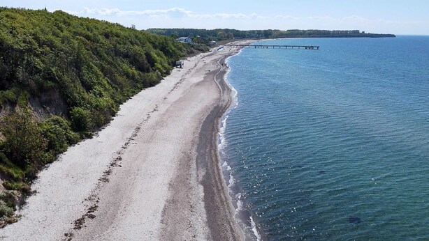 Uma estrada costeira com praia, um cais e uma colina arborizada.