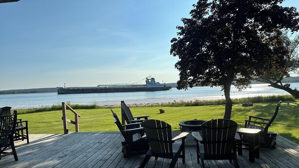 A wooden deck with Adirondack chairs and a table, overlooking a body of water with a large ship in the distance. - Freighters in front yard!  Private Sandy Beach - Large Deck - Best View on River (Sault Ste. Marie)