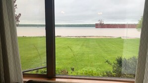 A room with a large window overlooking a grassy area and a body of water with a ship in the distance. - Freighters in front yard!  Private Sandy Beach - Large Deck - Best View on River (Sault Ste. Marie)