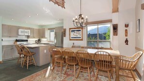 A spacious kitchen with a dining area, wooden chairs, a chandelier, and a view of mountains through the window.
