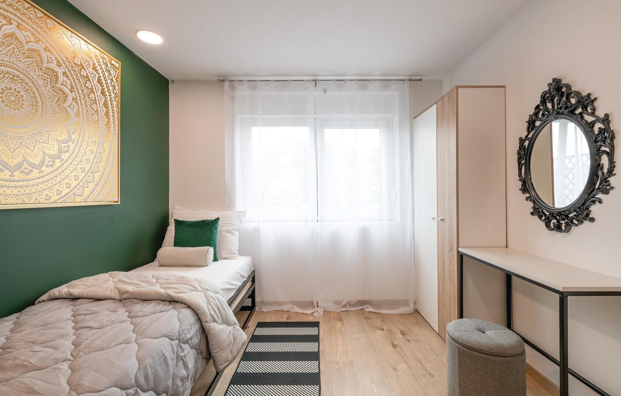 Un dormitorio con cama, mesita de noche, espejo y ventana con cortinas ligeras.