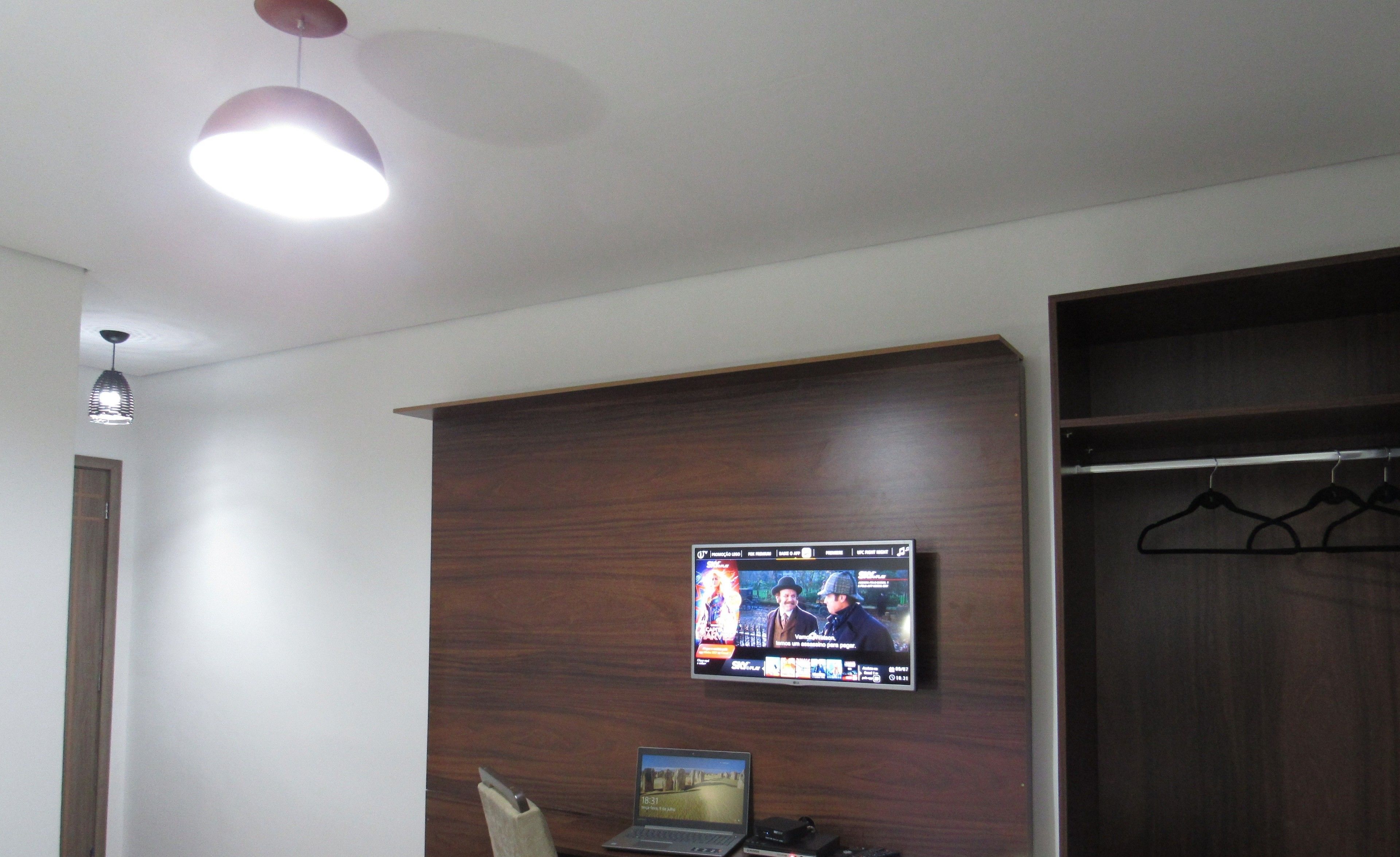 A room with a TV mounted on a wooden panel, a laptop on a desk, and a coat rack with hangers.
