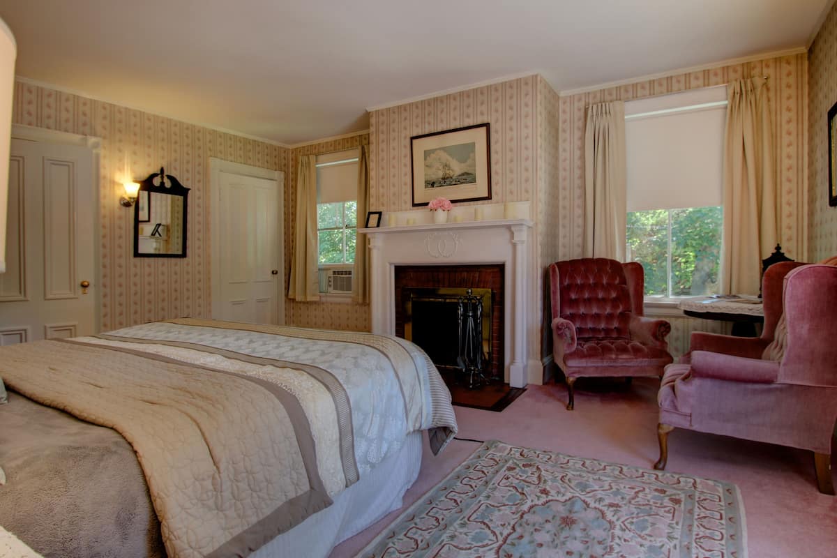 A bedroom with a large bed, a fireplace, a red armchair, and a window with curtains.