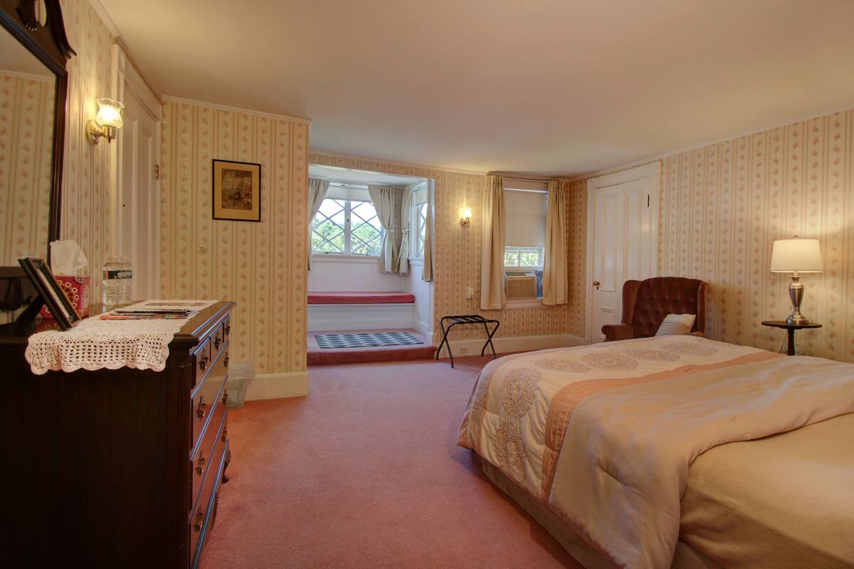 A hotel room with a bed, a chair, a desk with a lamp, a window with curtains, and a door.