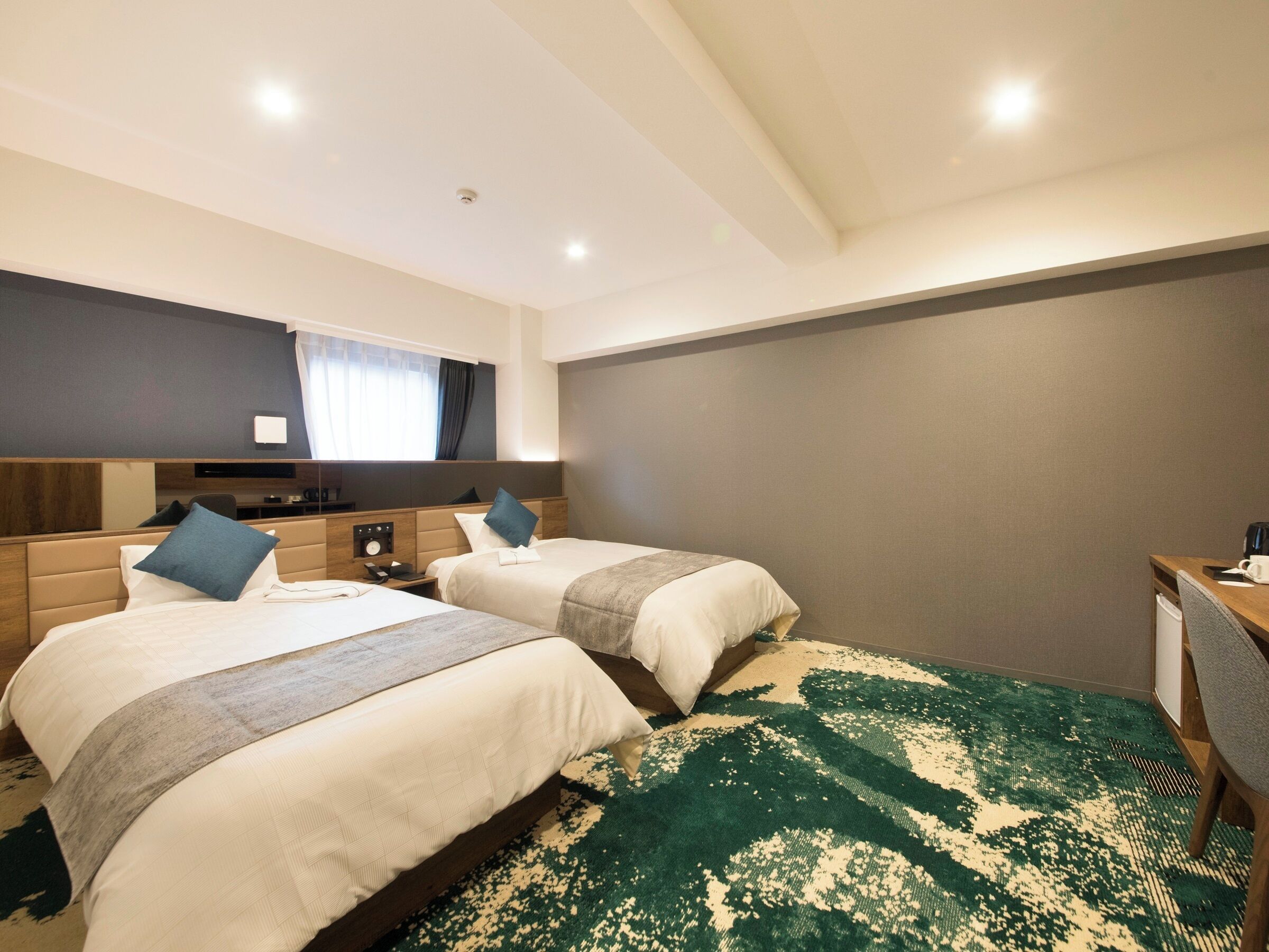Ein Hotelzimmer mit zwei Betten, einem Schreibtisch, einem Stuhl, einem Spiegel und einem großen Fenster mit Vorhängen.