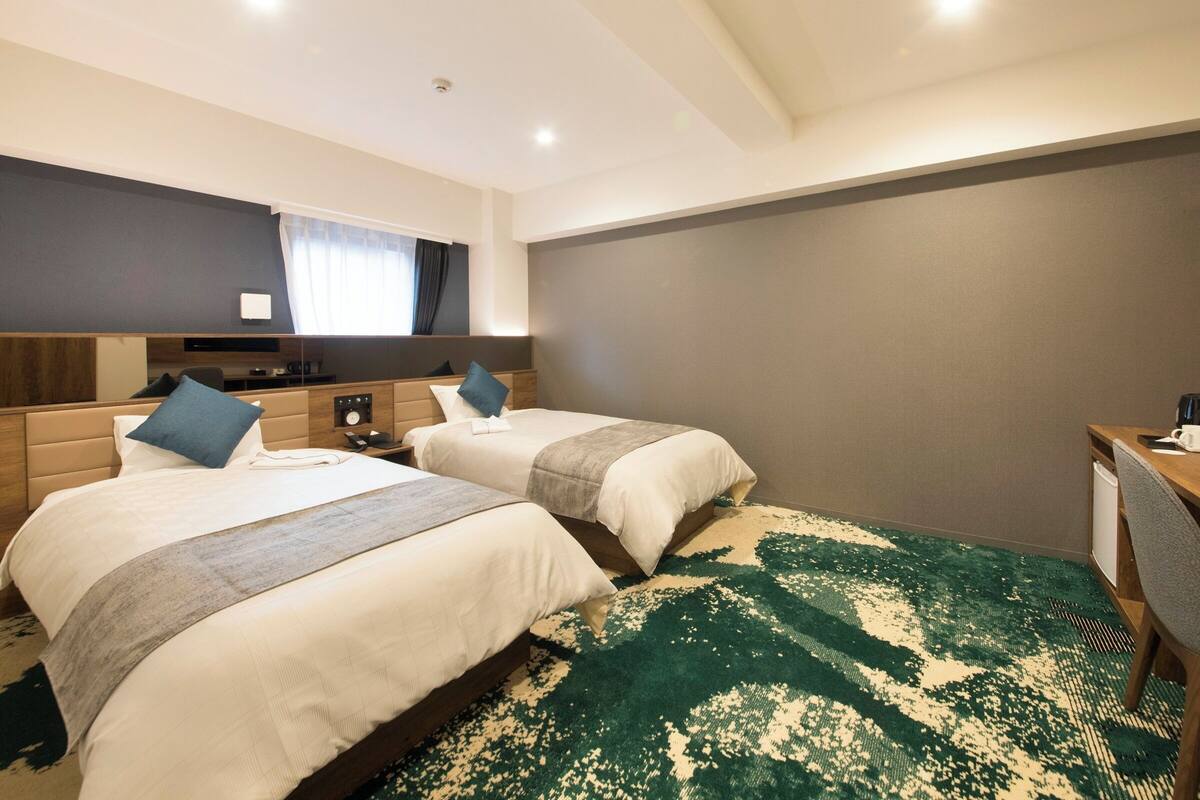 Ein Hotelzimmer mit zwei Betten, einem Schreibtisch, einem Stuhl, einem Spiegel und einem großen Fenster mit Vorhängen.