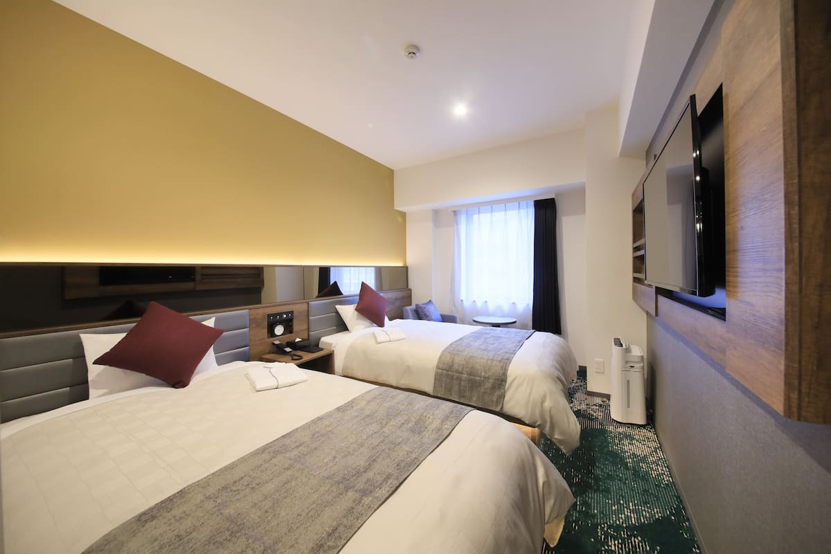 Ein Hotelzimmer mit zwei Betten, einem an der Wand montierten Fernseher, einem Nachttisch mit Lampe und einem Fenster mit Vorhängen.