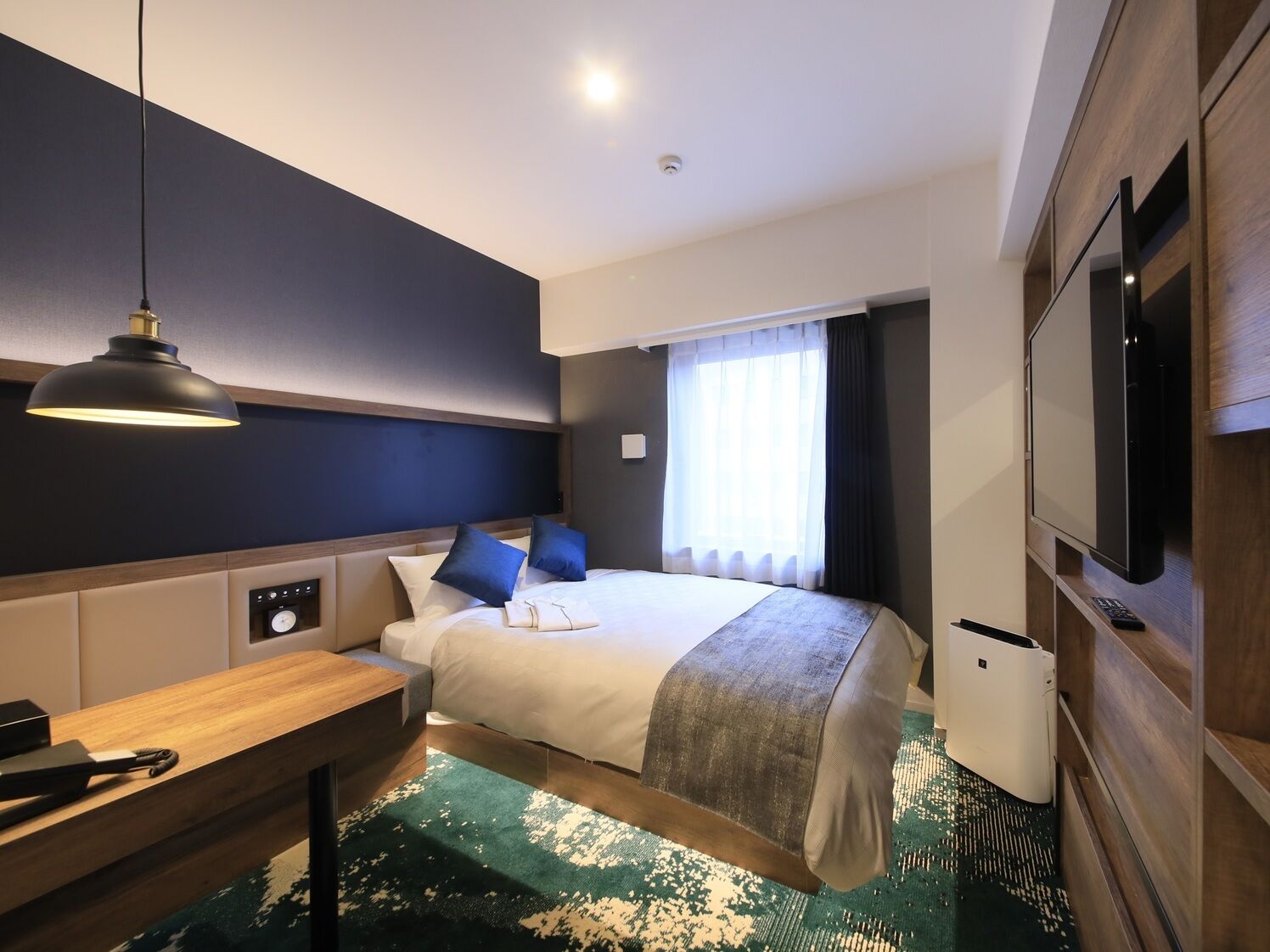 A hotel room with a bed, bedside table, lamp, TV, and a window with curtains.