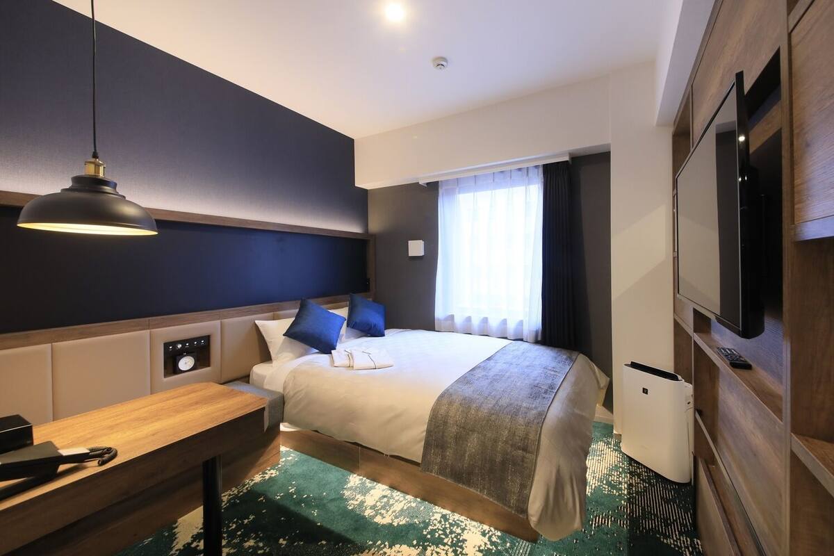 A hotel room with a bed, bedside table, lamp, TV, and a window with curtains.