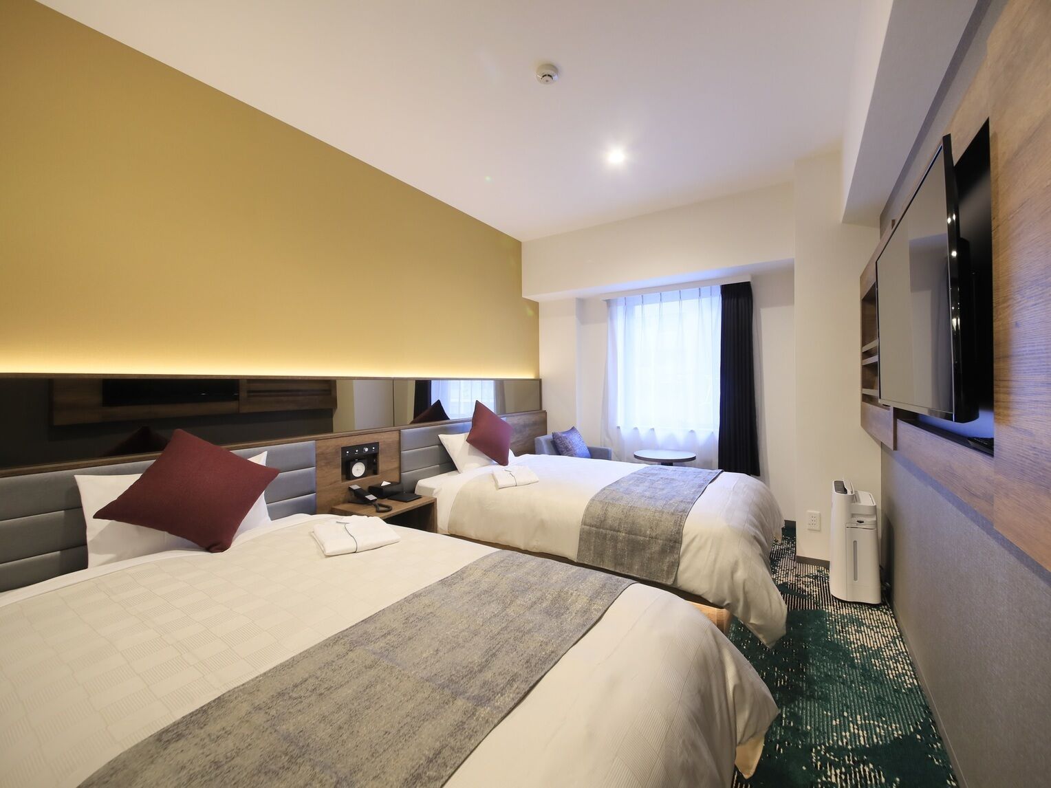 A hotel room with two beds, a flat-screen TV, a bedside table with a phone, a window with curtains, and a carpeted floor.
